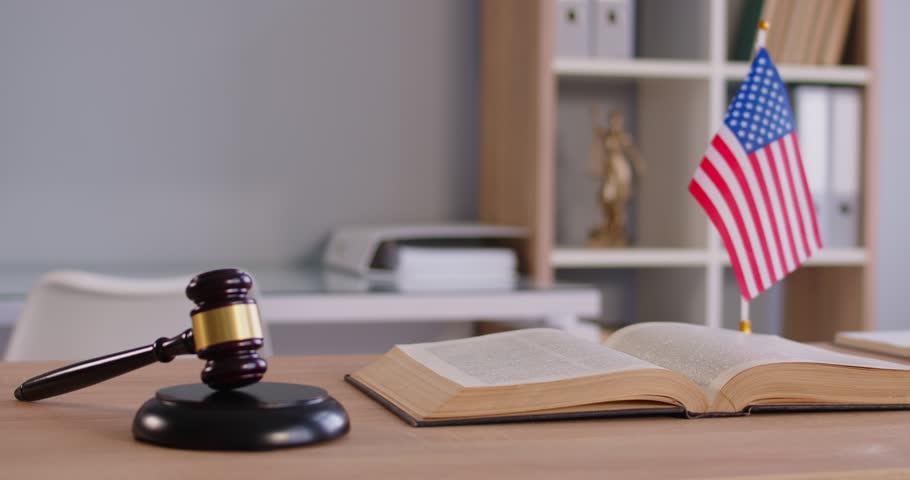 Americal legal system, prosecution and law. Judges gavel, law book and scales on table with Unites States of America flag in courtroom. Legal offiice with no people. Jusctice concept