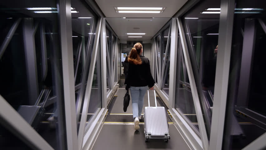 Confident businesswoman carry laptop bag and pulling wheeled suitcase while walking through airport jetway to airplane. Boarding time for night flight, slow motion shot, camera follow behind