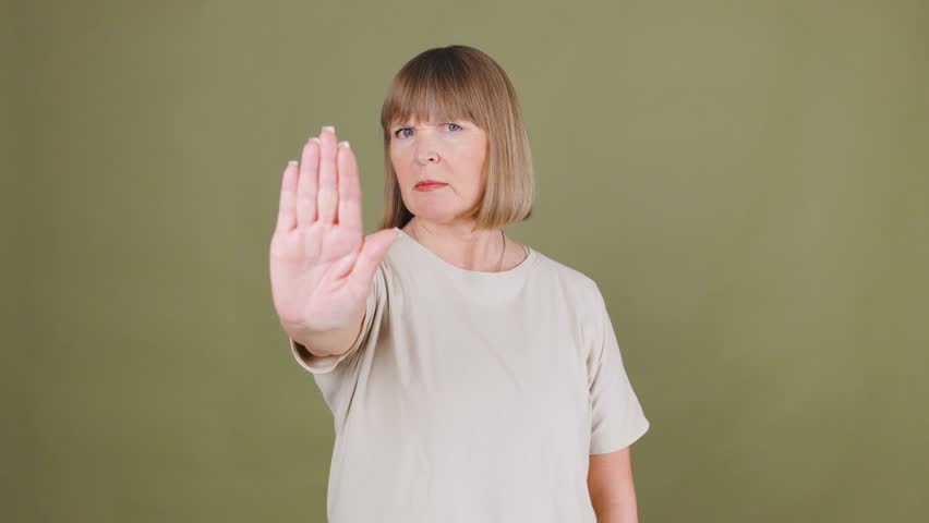 Senior woman signals to stop with raised hand, emphasizing a serious message of caution or refusal in a neutral setting