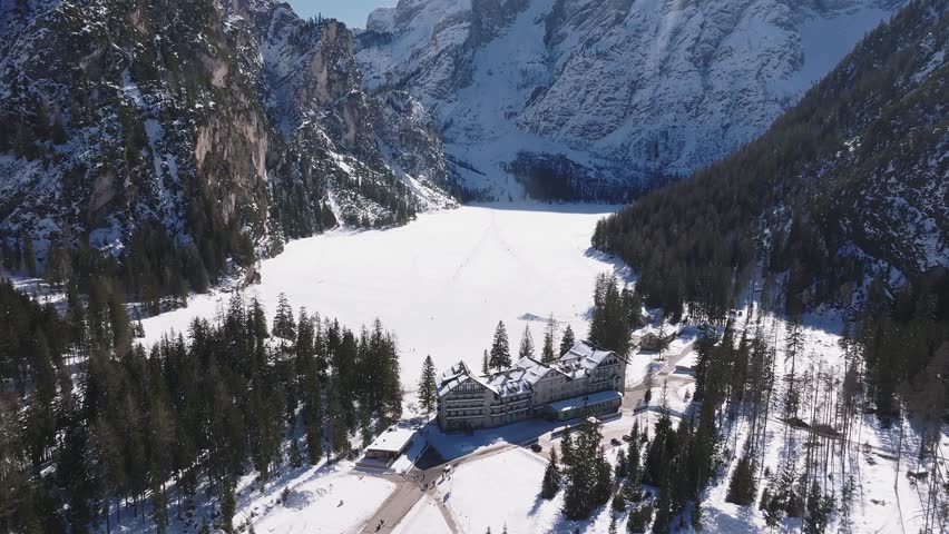 Aerial view of Braies Lake in northern Italy during winter, with a frozen surface, snow covered cliffs, evergreen forests, and scattered figures.