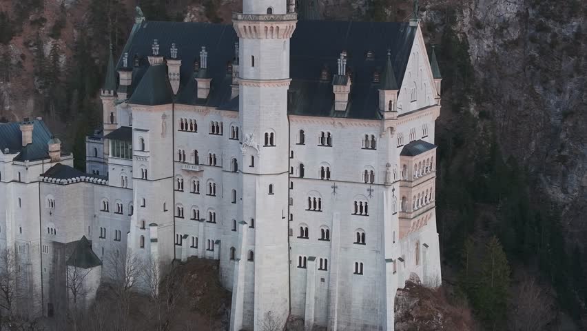 Aerial view of Neuschwanstein Castle in Bavaria, Germany, at sunset. The video pans around the castle, showing mountains, forest, and a distant church.