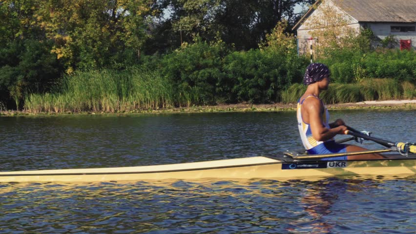 Rowing team training. Side view of 2 young caucasian male rowers, during a rowing practice, athlete sitting in a boat in the river Dnipro, rows through a calm water sunny day, autumn. 4k footage