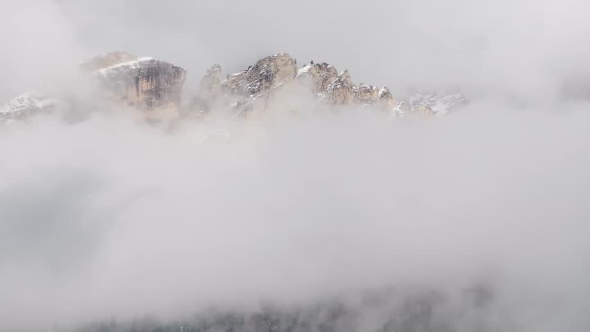 Aerial view of the Dolomite mountains with snow dusted peaks emerging through shifting clouds. The camera sweeps slowly, revealing the rugged terrain.