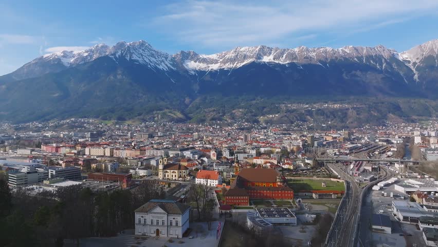 Aerial pan of Innsbruck, Austria, featuring the Nordkette mountains, a red roofed building with a helipad, and a mix of urban architecture below.