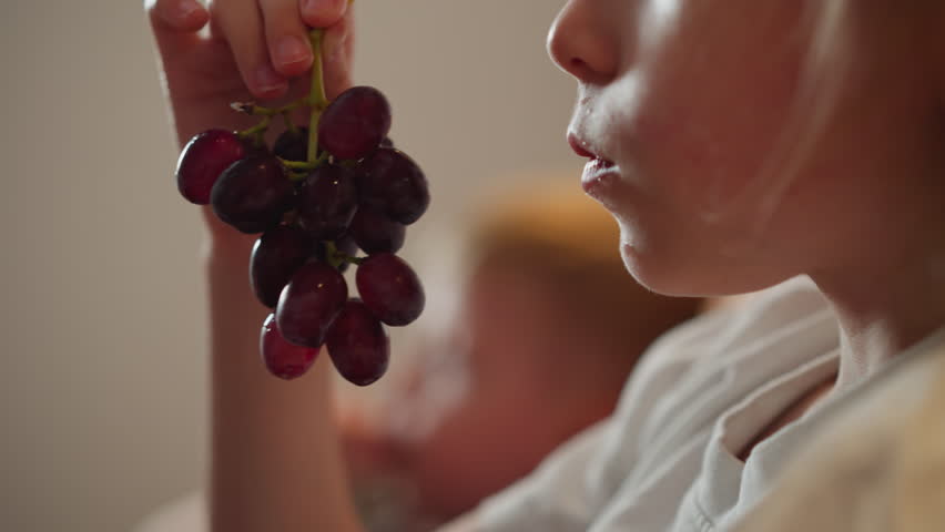 close up of white girl holding bunch of grapes and eating with intense focus while blurred figure appears in background creating warm peaceful childhood