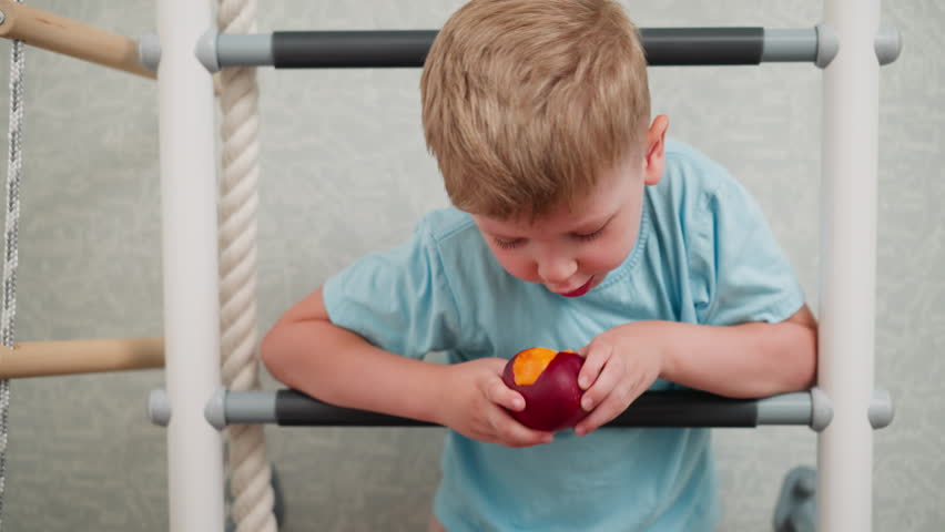 Young boy leans over indoor ladder munching on ripe mango with focus on juicy fruit and playful child moment in bright room showcasing fun and healthy snacking during casual home activity