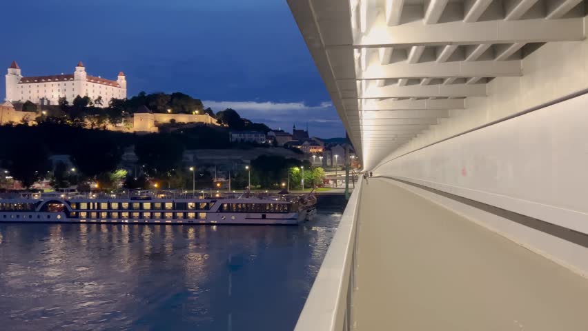 A scenic night view of Bratislava Castle illuminated atop a hill, overlooking the Danube River. The reflections of city lights shimmer on the water, with boats docked along the riverbank.