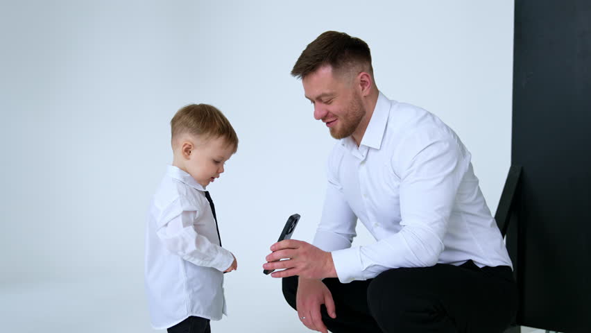 Proud father in white shirt and his son in white shirt and black tie. Father shows a smartphone to his baby boy.