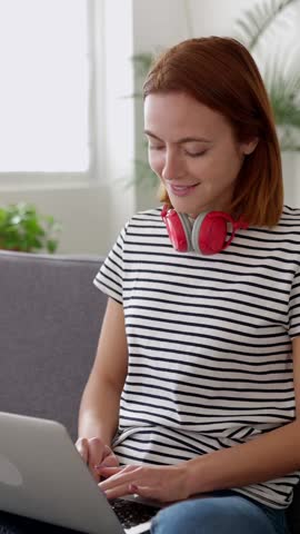 Vertical HD video of young adult woman using a laptop computer while studying online using laptop computer sitting on sofa at modern apartment. Education and technology concept.