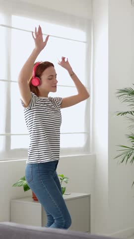 Vertical HD video of joyful young woman listening to music with wireless headphones and dancing in the living room, taking a break from work or studies. Happy people and carefree concept.