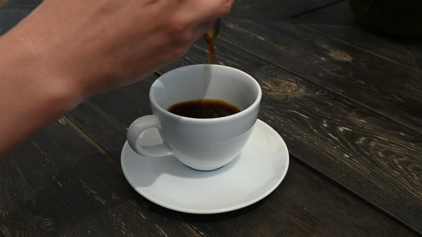 White cup receiving freshly brewed dark coffee on wooden surface, lifting by hand with careful, warm beverage gesture showcasing morning relaxation moment