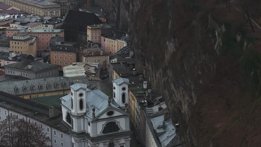 Aerial view of Salzburg, Austria, featuring Hohensalzburg Fortress, urban architecture, wooded hills, and distant mountains under an overcast sky.