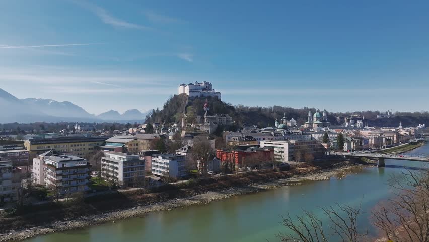 Aerial view of Salzburg, Austria, showing the Salzach River, Hohensalzburg Fortress, Salzburg Cathedral, bridges, and tree lined pathways.