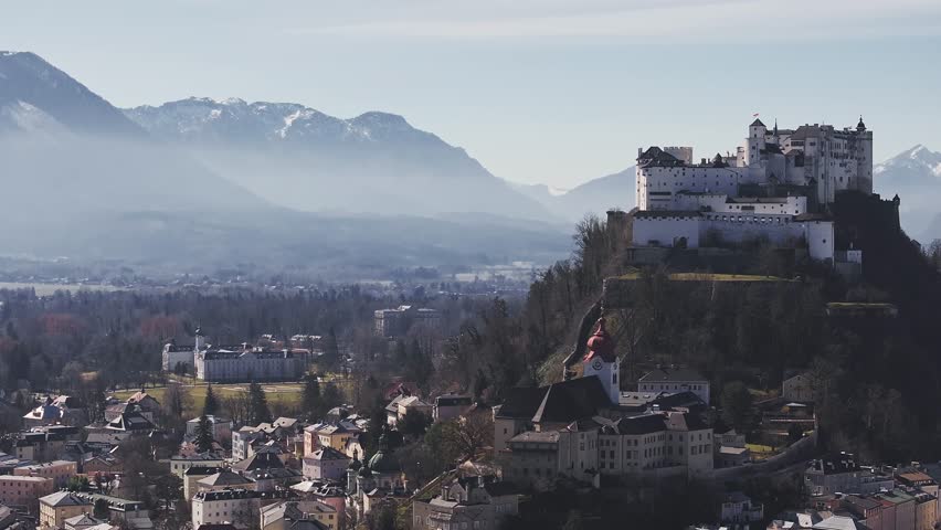 Aerial view of Salzburg, Austria, featuring Hohensalzburg Fortress on a hill, historic architecture, and the Alps with a rugged mountain peak in motion.