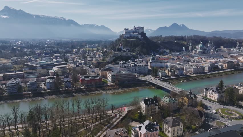 Aerial view of Salzburg, Austria, featuring the Salzach River, Hohensalzburg Fortress, historic architecture, and surrounding mountains. Camera pans smoothly.