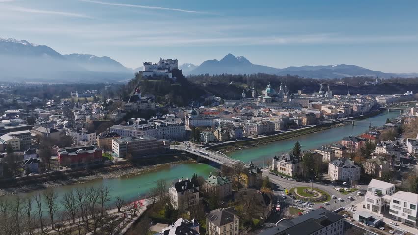 Aerial pan of Salzburg, Austria, featuring Hohensalzburg Fortress, the Salzach River, historic architecture, and snow capped mountains in the background.