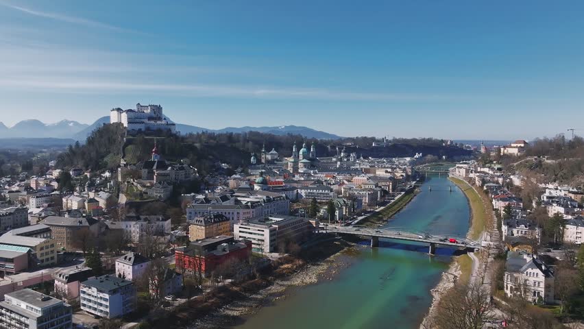 Smooth ascending drone view of Salzburg, Austria, featuring Hohensalzburg Fortress, the Salzach River, tree lined pathways, and urban greenery.