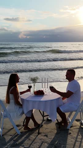 A lovely couple sits together enjoying a romantic beach dinner at sunset, joyfully toasting with their glasses of wine