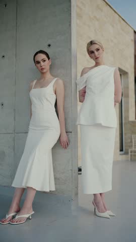 Two fashionable women in stylish white dresses posing outdoors against a concrete wall. Modern and minimalist setting highlighting elegant fashion and contemporary styles.