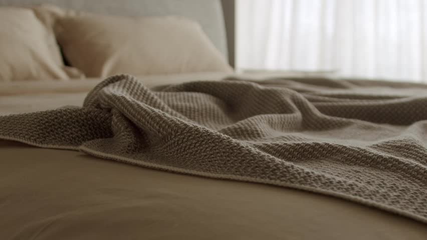 A cozy and serene bedroom featuring a beige knit blanket spread on a soft bed under natural sunlight, creating a warm and inviting atmosphere. Cozy Bed with Beige Knit Blanket in Serene,Sunlit Bedroom
