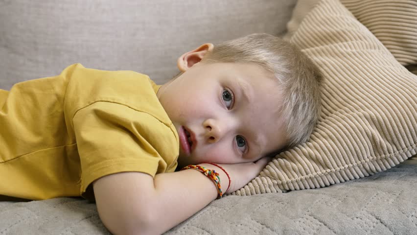 Blond young male child lying on comfortable sofa, head resting on pillow, wearing yellow t-shirt and bracelet, displaying bored, pensive expression with melancholic undertones