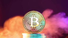 Bitcoin cryptocurrency coin standing on a stack of coins surrounded by colorful smoke on a dark background, representing the volatility and uncertainty of the cryptocurrency market - Powered by Shutterstock - Get 15% off with code: PIKWIZARD15