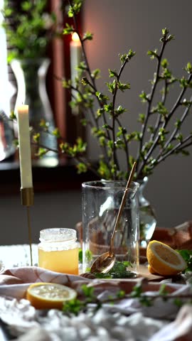 A person pours hot water from a glass teapot into a tall glass containing green leaves, likely currant leaves, and other herbs. Tea with currant leaves. Food photo, candles, cozy tea party