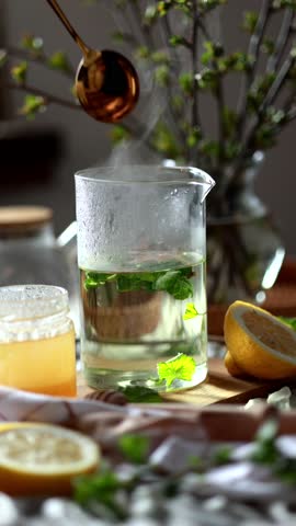A person stirring tea with a golden spoon into a tall glass containing green leaves, likely currant leaves, and other herbs. Tea with currant leaves. Food photo, candles, cozy tea party
