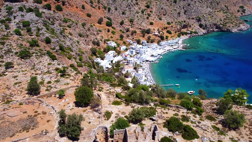 High drone shot of Loutro village and beach from the side. White buildings contrast with blue-green sea, surrounded by mountains on the southern coast of Crete.