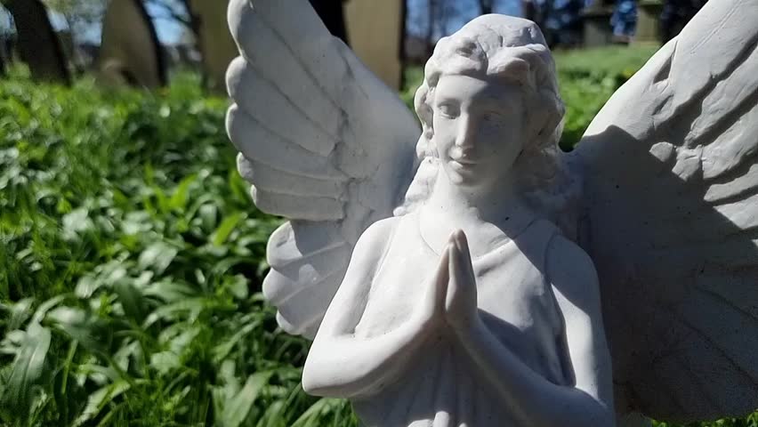 Small sunlit marble gravestone angel memorial with wings praying close up against summer foliage