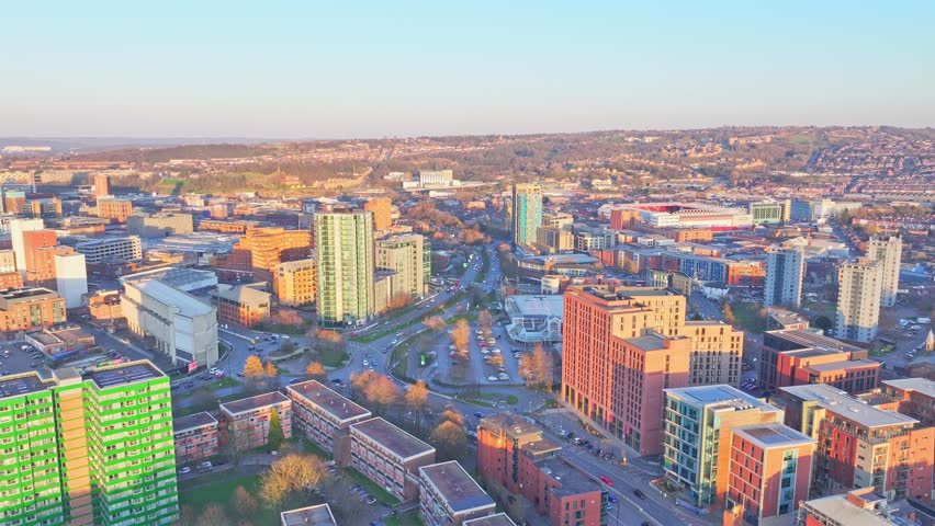 The drone descends slowly in morning light over Collegiate Crescent, Sheffield. Buildings, a football stadium, autumn trees, traffic, and long shadows create an expansive view of the urban landscape.