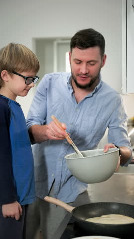 Father teaches his son how to properly pour batter into a heated frying pan. Medium long shot. Slow motion