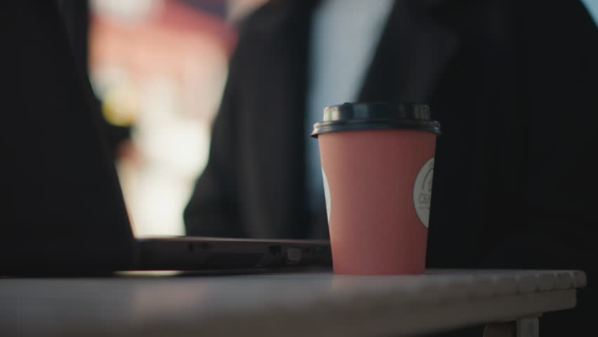 Close-up of laptop on table with hand in black coat, polished nails, dropping a pink coffee cup onto table surface sits down, blurred background