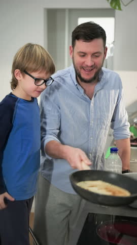 The boy whaches how his father shake and flip a pancake in the air on a hot frying pan. Medium long shot. Slow motion.