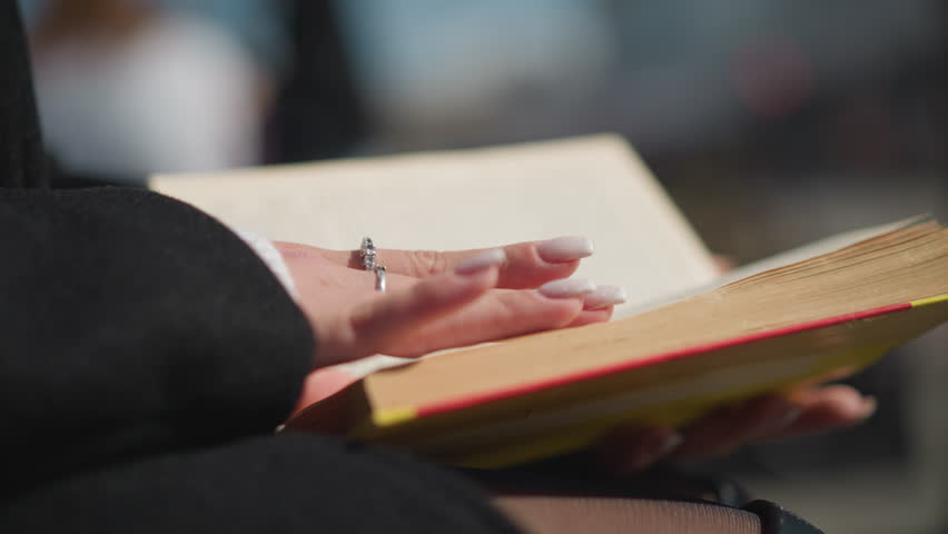 Close-up of a woman with manicured nails gently flipping through the pages of a book, revealing a vibrant edge with sunlight reflecting off her hand and the surrounding soft background