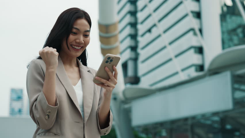 Portrait of Thai businesswoman happy of unexpected financial bonus against office building. Asian lady checks mobile banking app outdoors. Female manager celebrates victory
