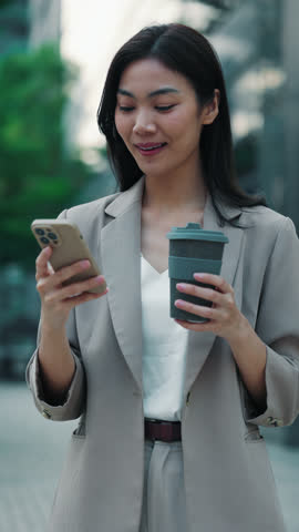 Portrait of Thai businesswoman with coffee in reusable cup typing message on smartphone while standing on street. Asian woman engages in online communication with clients using phone. Vertical video