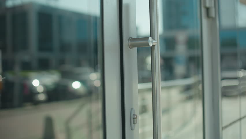 Close up of professional woman with handbag opening glass door stepping out of building, focus on hand gripping metal handle, reflections visible on door, modern urban setting with sleek architecture