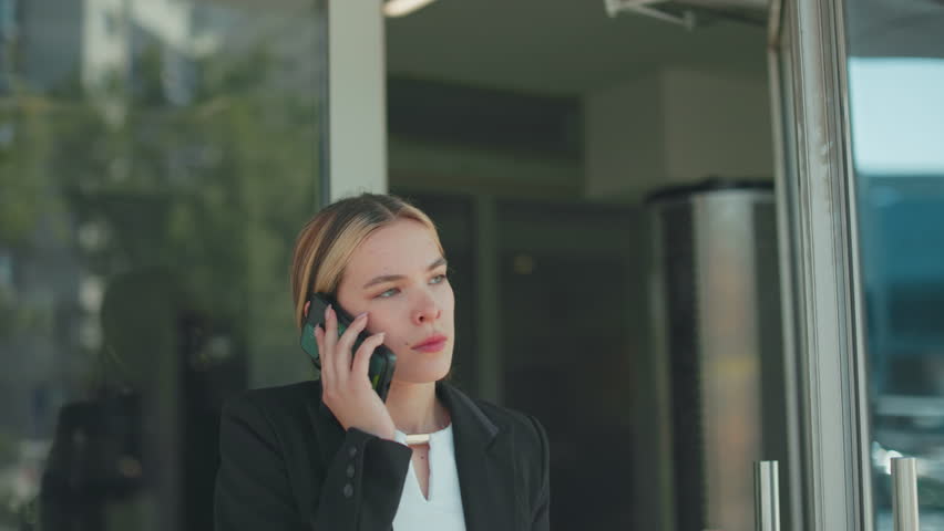 Young lady frustrated during phone call after work, about to lower phone while exiting office building, dressed in formal outfit, glass door reflects urban setting with vehicles in background
