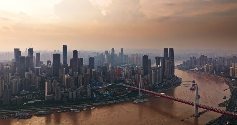 Aerial view presenting a wide panorama of the Chongqing skyline and bridge along the river under a hazy sky.