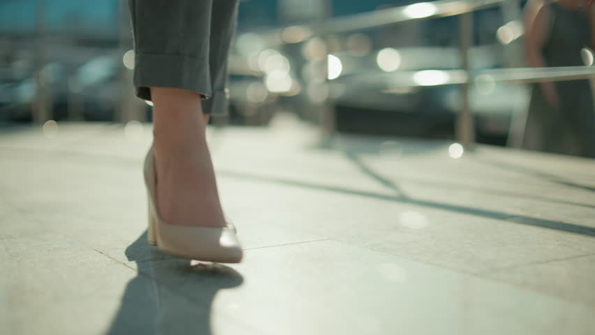 Close up leg view of woman wearing heels walking confidently along iron railing with sunlight reflecting on pavement, blurred parked cars and pedestrians in background