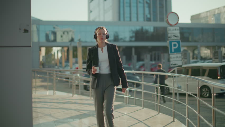 Business lady in headphones enjoying music outdoors holding coffee cup with relaxed expression, sunlight casting warm glow in urban setting with office buildings, parked cars, and metal railings