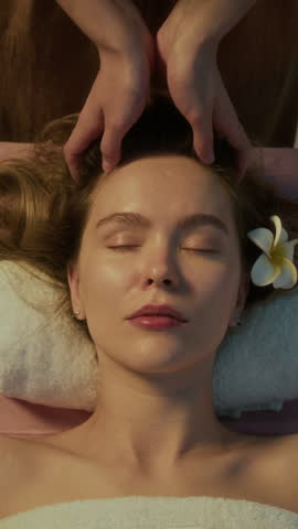Vertical top view portrait shot of happy woman smiling at looking at camera while receiving relaxing head massage in spa