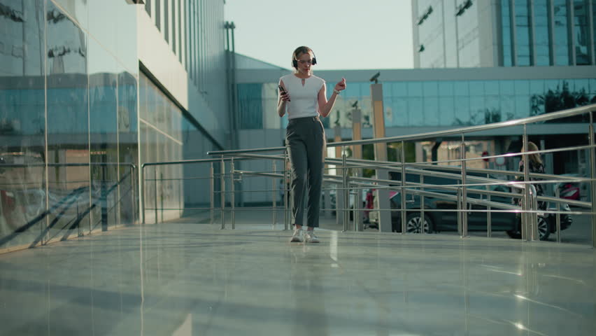 Young lady dancing with phone and headphones on urban walkway, moving rhythmically near modern buildings with reflective glass, parked cars, visible signage, and another woman walking in background