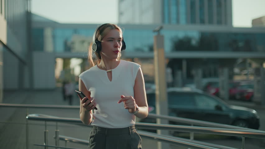 White woman enjoying music with headphones holding phone, sunlight illuminating face and glass building reflecting urban surroundings and hair clipped back