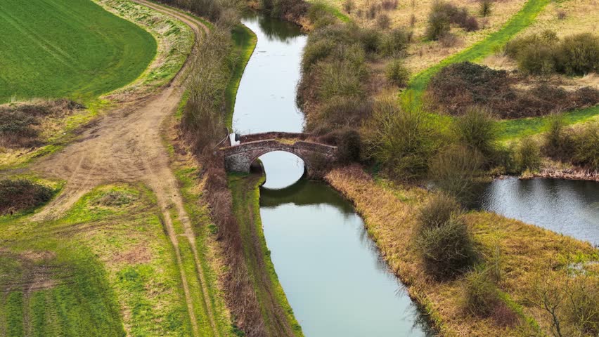 Ancient brick bridge arching over historic canal in United Kingdom. Still water reflecting old masonry structure amid green countryside. Narrow waterway cutting through rural landscape with dirt paths