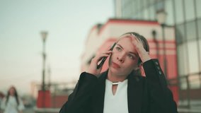 Frustrated white lady in professional outfit expressing exhaustion while on phone call in urban environment, hand raised in stress, people blurred in distant background - Powered by Shutterstock - Get 15% off with code: PIKWIZARD15