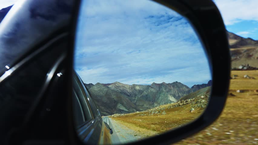 As I am driving through peaceful and scenic roads, I admire the stunning reflections of the mountainous landscape seen in the side mirror of my vehicle, capturing the beauty of my journey perfectly
