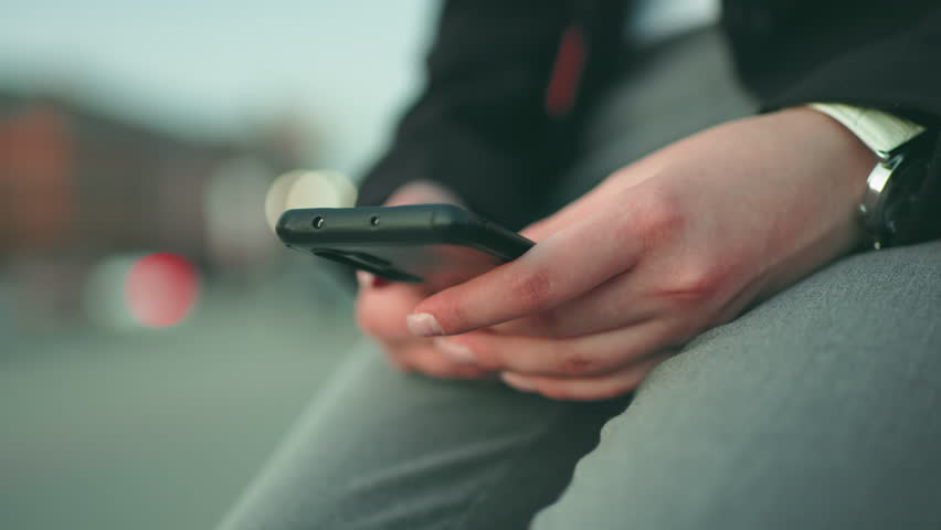 Close up soft focus of young lady with artificial nails operating phone in both hands while seated, wearing black jacket and wristwatch, with soft bokeh lights and blurred background