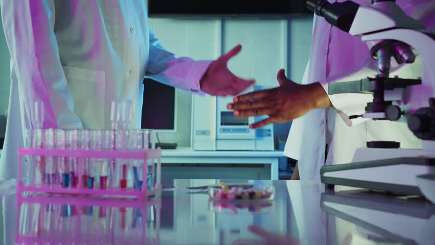 Two scientists in lab coats shaking hands over laboratory table with test tubes and microscope, celebrating a successful experiment or research collaboration